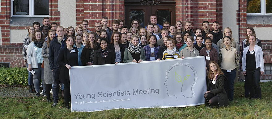 Gruppenfoto des Young-Scientist Meeting 2022. Eine Gruppe mit Menschen steht vor Gebäude aus Backstein und hält ein Transparent mit Logo der Veranstaltung hoch.