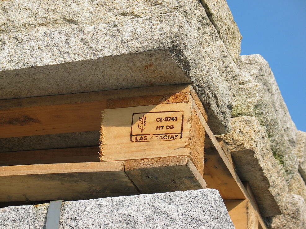 Eine Holzpalette, auf der Steine lagern, ist mit einem Brandzeichen versehen. Das Zeichen belegt, dass die Palette thermisch behandelt wurde. Auf diese Weise soll verhindert werden, dass mit dem Holz Pflanzenschädlinge verschleppt werden..