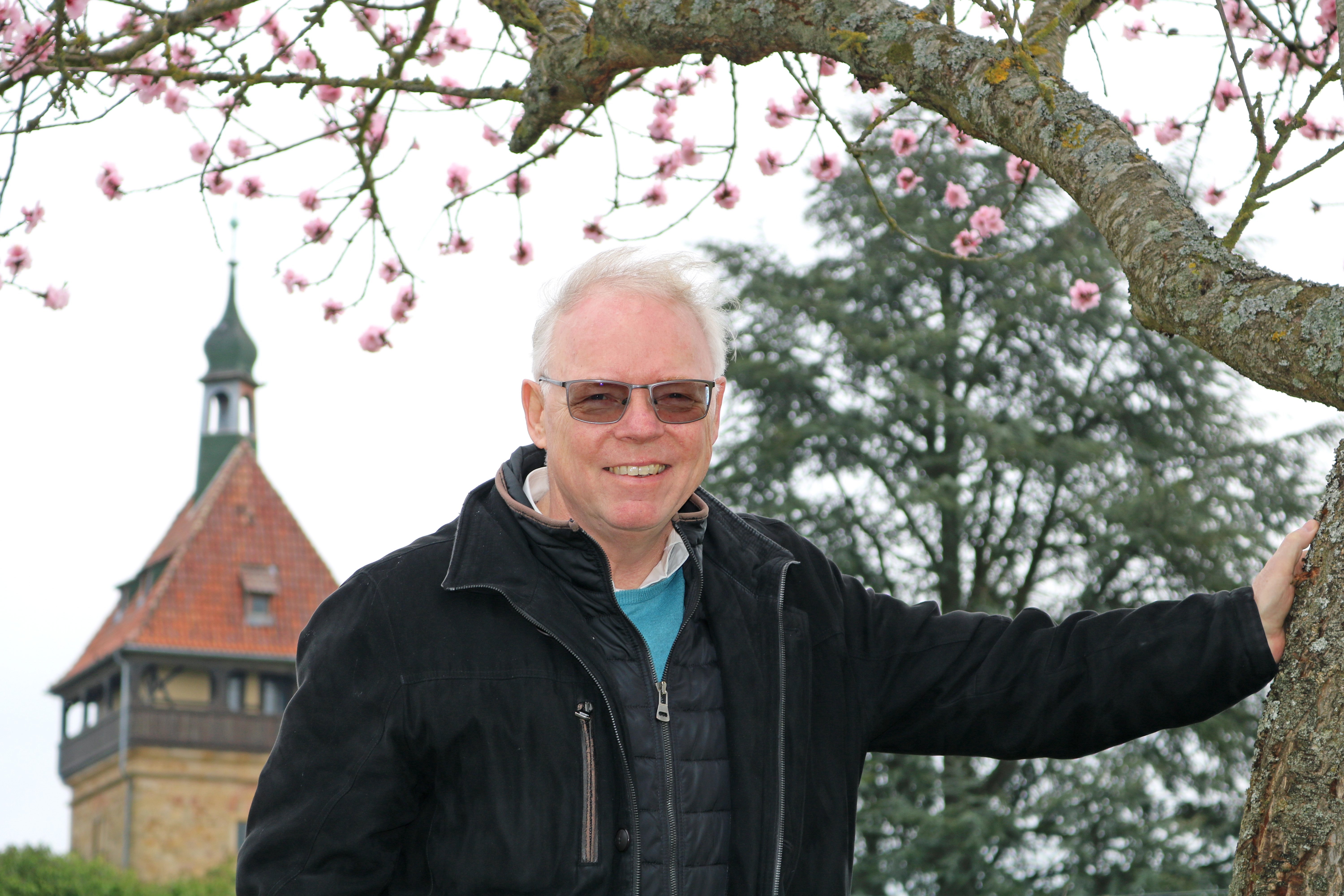 Nahaufnahme eines älteren grauhaarigen Mannes mit getönter Brille, der sich gegen einen blühenden Baum stützt. Im Hintergrund ist ein alter Turm zu sehen.