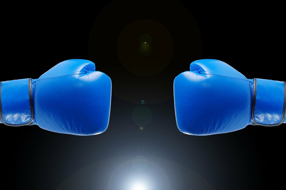 Zwei geballte Fäuste in blauen Boxhandschuhen erscheinen aus den gegenüberliegenden vertikalen Bildrand vor schwarzem Hintergrund. Sie schweben voreinander. Konfrontation wird angedeutet.