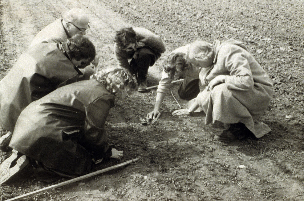 Altes Sepia-foto: Mehrere Menschen knien auf einer Ackerfläche und schauen konzentriert auf einen Punkt.