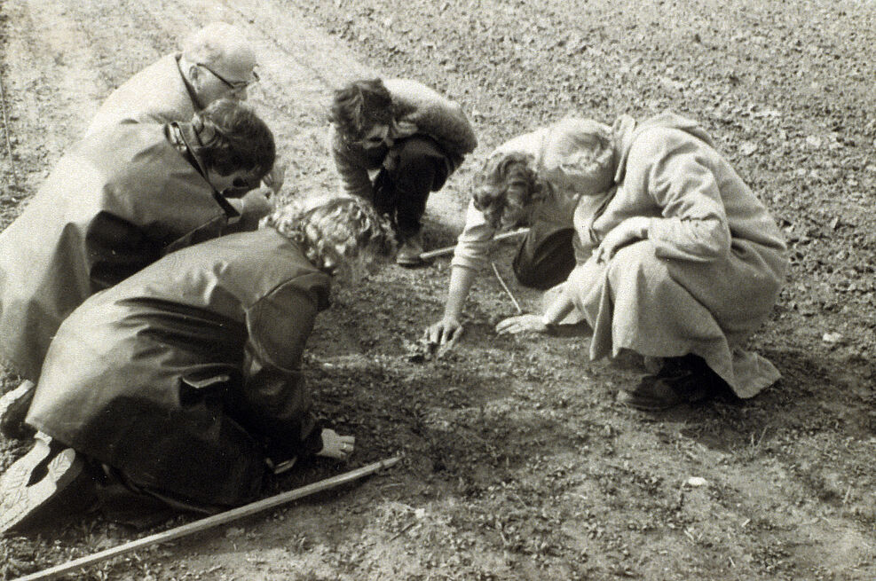 
	Altes Sepia-foto: Mehrere Menschen knien auf einer Ackerfläche und schauen konzentriert auf einen Punkt.
