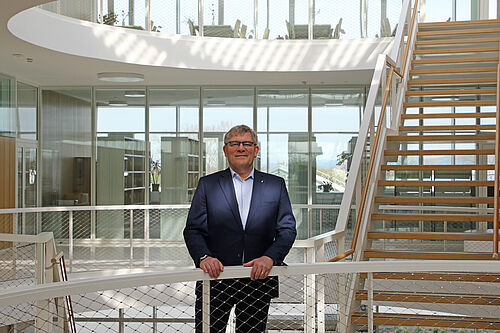 Älterer Mann im Anzug steht vor einem Geländer in einem hellem Foyer. Tageslicht wirft Muster auf Treppen und Wände hinter ihm.