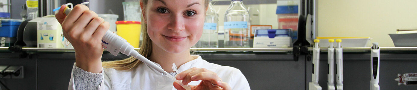 Ein junges Mädchen guckt in die Kamera und pipettiert dabei Flüssigkeit in ein Röhrchen. Im Hintergrund: Laborumfeld.