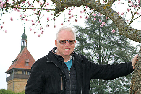 Nahaufnahme eines älteren grauhaarigen Mannes mit getönter Brille, der sich gegen einen blühenden Baum stützt. Im Hintergrund ist ein alter Turm zu sehen.