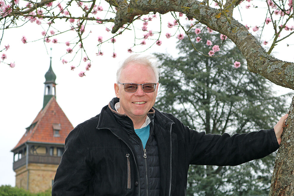 
	Nahaufnahme eines älteren grauhaarigen Mannes mit getönter Brille, der sich gegen einen blühenden Baum stützt. Im Hintergrund ist ein alter Turm zu sehen.
